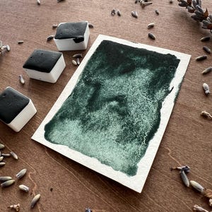 May include: A still-life composition featuring watercolor paints and dried lavender on a wooden surface. Three square watercolor paint blocks with black paint are next to a rectangular watercolor swatch in shades of green. Dried lavender buds and sprigs are scattered around the objects.