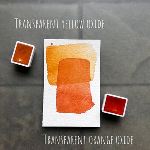 May include: Two watercolor paint pans, one orange and one yellow, with a painted swatch of each color on a white card. The card is labeled "TRANSPARENT YELLOW OXIDE" and "TRANSPARENT ORANGE OXIDE".