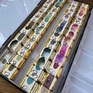 K&ouml;nnte beinhalten: Eine offene Metall-Aquarellfarbenbox mit einzeln verpackten Aquarellfarbenstiften. Jeder Stift hat eine goldene Verpackung und ein Aquarelldesign auf dem Etikett. Die Farbpalette umfasst Gr&uuml;nt&ouml;ne, Blau, Lila und Braun.