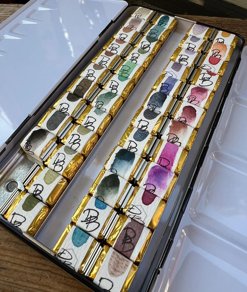 K&ouml;nnte beinhalten: Eine offene Metall-Aquarellfarbenbox mit einzeln verpackten Aquarellfarbenstiften. Jeder Stift hat eine goldene Verpackung und ein Aquarelldesign auf dem Etikett. Die Farbpalette umfasst Gr&uuml;nt&ouml;ne, Blau, Lila und Braun.