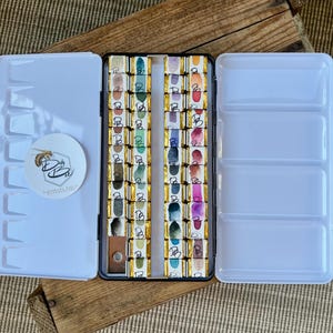K&ouml;nnte beinhalten: Eine wei&szlig;e Aquarellfarbpalette mit offenem Deckel, die Reihen farbenfroher Farbkuchen zeigt. Die Palette enth&auml;lt einen runden Aufkleber mit einem Bienenlogo und dem Text "D by Ba". Die Palette befindet sich auf einer Holzoberfl&auml;che.