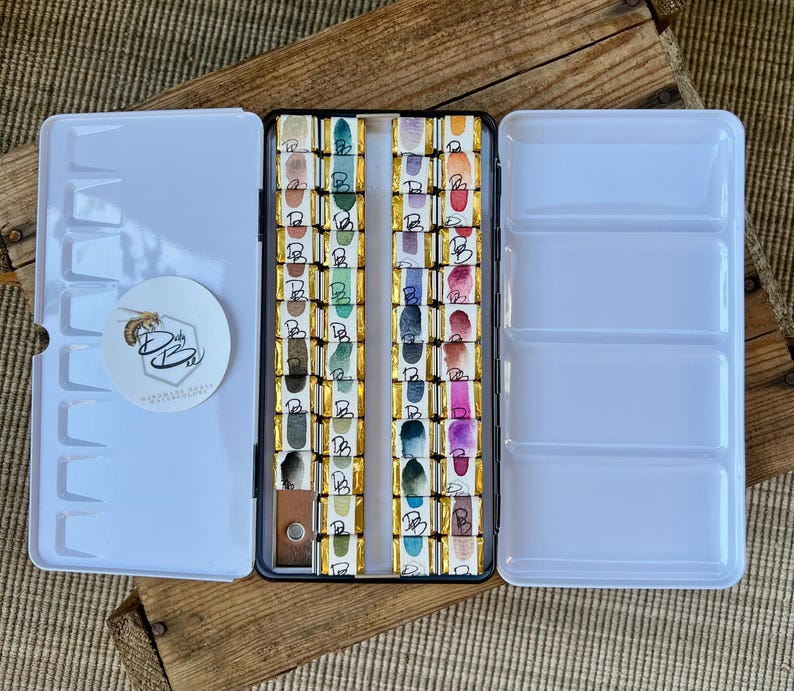 K&ouml;nnte beinhalten: Eine wei&szlig;e Aquarellfarbpalette mit offenem Deckel, die Reihen farbenfroher Farbkuchen zeigt. Die Palette enth&auml;lt einen runden Aufkleber mit einem Bienenlogo und dem Text "D by Ba". Die Palette befindet sich auf einer Holzoberfl&auml;che.