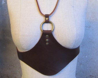 May include: A brown leather chest harness with a circular brass ring and three small brass rivets. The harness is displayed on a mannequin. The neck strap is a combination of brown and tan leather.