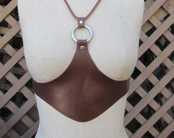 May include: A brown leather halter top with a silver ring and a brown leather strap. The halter top is on a white mannequin.