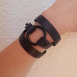 May include: A black leather bracelet with a large, round, black metal ring. The bracelet is wrapped around a wrist.