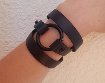 May include: A black leather bracelet with a large, round, black metal ring. The bracelet is wrapped around a wrist.