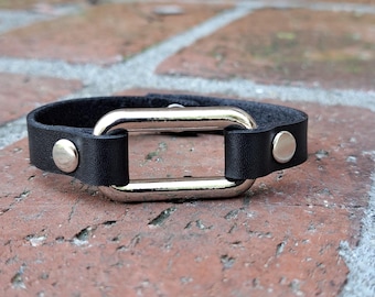 May include: A black leather bracelet with a silver oval buckle. The bracelet has two silver studs on either side of the buckle.