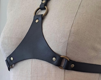 May include: Black leather body harness with brass-colored metal rings and rivets. The harness features a triangular chest piece and adjustable straps. The design is simple and elegant, suitable for various styles.