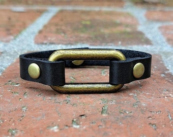 May include: A black leather bracelet with a gold-toned oval clasp. The bracelet has two gold-toned rivets on each side of the clasp.