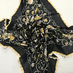 May include: A black sheer scarf with a floral pattern of yellow and white flowers. The scarf has a crocheted border with white and yellow yarn.
