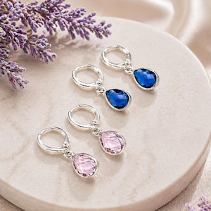May include: Silver hoop earrings with teardrop-shaped blue and pink gemstones. The earrings are displayed on a white circular surface, with sprigs of lavender in the background. The gemstones have a faceted cut, reflecting light.