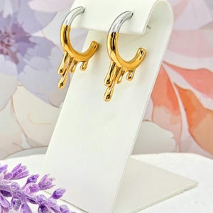 May include: A pair of gold-tone hoop earrings with a silver-tone top section. The earrings have a unique design with a dripping effect, adding a modern touch. They are displayed on a white stand against a floral background.