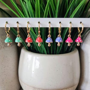 May include: A collection of seven gold hoop earrings, each featuring a mushroom charm. The mushroom caps are in various colors: green, white, red, purple, and pink, all with white polka dots. The earrings are displayed against a backdrop of green plants.
