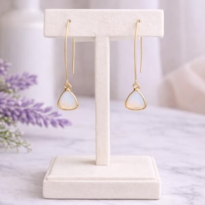 May include: A pair of gold-toned drop earrings with a triangular, opalescent stone. The earrings are displayed on a white jewelry stand, showcasing their delicate design. The earrings have a minimalist style, perfect for everyday wear.