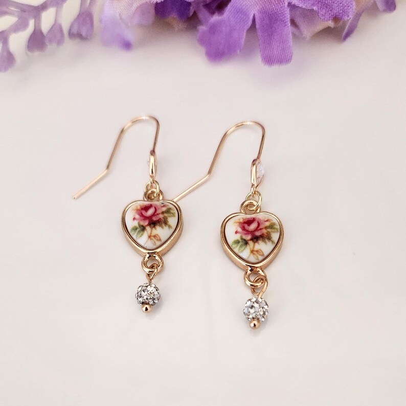 May include: Gold heart-shaped earrings with a floral design and a small crystal dangle. The floral design features a pink rose on a white background.