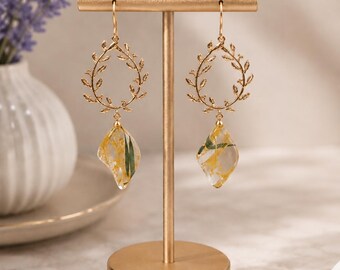 May include: A pair of gold-toned drop earrings featuring a circular frame shaped like a leafy vine. Suspended from the bottom of each vine is a clear, diamond-shaped resin pendant containing embedded yellow flower petals and green foliage.