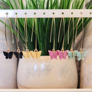 May include: A collection of butterfly dangle earrings in various colors, including black, yellow, pink, and teal. The earrings are displayed on a white rack against a backdrop of green foliage. Each earring features a gold-colored wire.