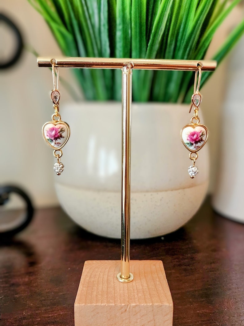 May include: Gold heart-shaped earrings with a pink rose floral design and clear crystal accents.