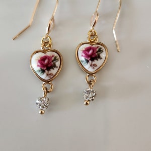 May include: Gold heart-shaped earrings with a white porcelain rose design and a clear crystal dangle.