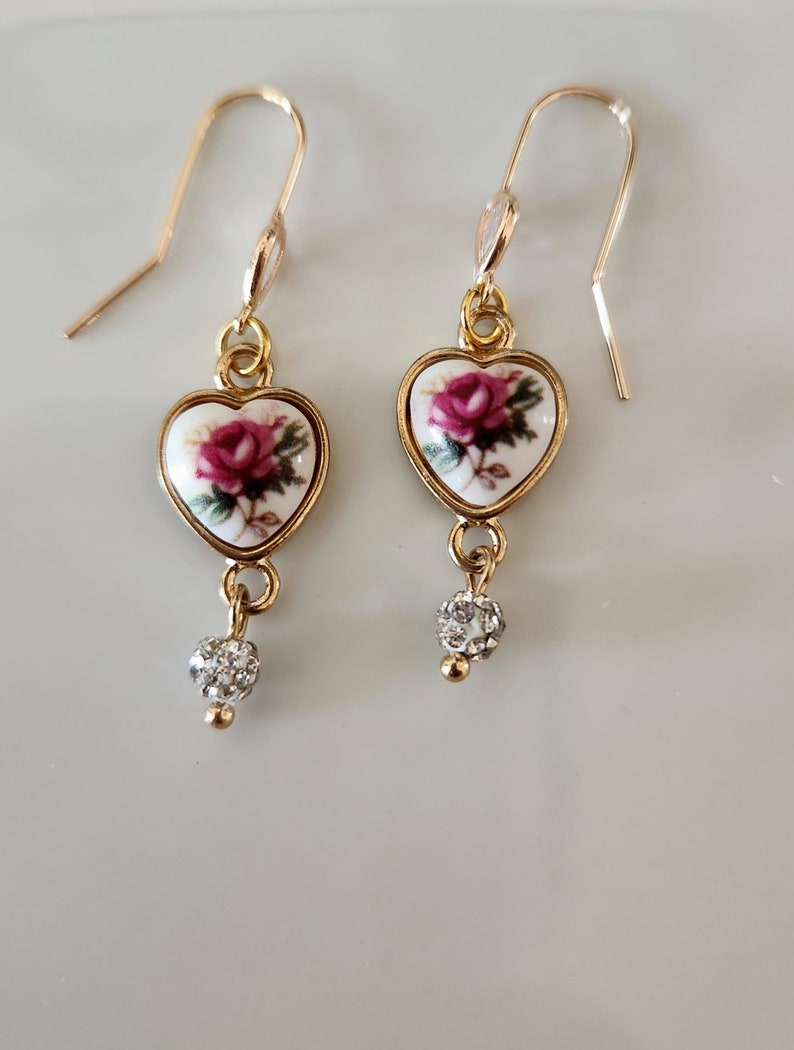 May include: Gold heart-shaped earrings with a white porcelain rose design and a clear crystal dangle.