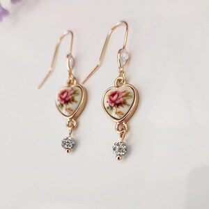 May include: Gold heart-shaped earrings with a floral design and a small crystal dangle. The floral design features a pink rose on a white background.