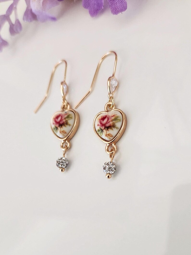 May include: Gold heart-shaped earrings with a floral design and a small crystal dangle. The floral design features a pink rose on a white background.