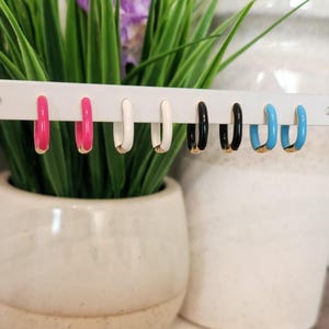 May include: Six pairs of small hoop earrings with colorful enamel. The hoops are gold-toned and have a thin, rounded shape. The colors are pink, white, black, and blue.