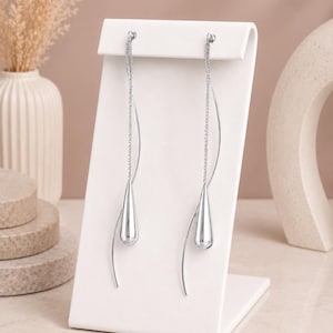 May include: A pair of long silver-tone earrings featuring a delicate chain and a smooth, polished teardrop-shaped pendant. Each earring includes a slender, curved metal wire that extends downward, creating a sleek and elongated silhouette.
