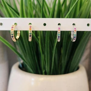 May include: Four hoop earrings with colorful gemstones are displayed on a white bar. The earrings are gold and silver, each adorned with a row of rainbow-colored stones. The earrings are in front of a green plant in a white pot.