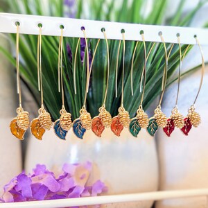 May include: A collection of gold-toned dangle earrings. Each earring features a small, textured pinecone charm and a colorful leaf charm in shades of yellow, blue, red, and green. The earrings are displayed on a white rack against a backdrop of greenery.
