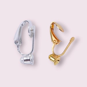 May include: Two earring clip-on findings, one silver-colored and one gold-colored. The silver clip-on has a curved top and a cylindrical base. The gold clip-on has a hinged top and a curved lever. Both are designed for jewelry making.