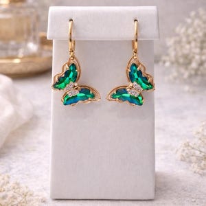 May include: A pair of gold-toned butterfly earrings with emerald green and clear crystal accents. The butterfly wings are filled with green crystals, and the earrings have a delicate, elegant design. The earrings are displayed on a white stand.