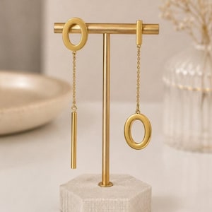 May include: A pair of asymmetrical gold-toned earrings displayed on a T-shaped stand. One earring features an oval hoop with a dangling bar, while the other features a rectangular bar with a dangling oval hoop, both connected by delicate gold chains.