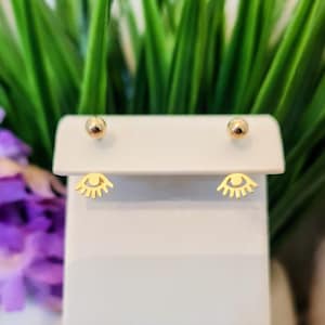 May include: Gold-colored stud earrings. One pair features a small, round stud and a stylized eye design with eyelashes. The earrings are displayed on a white surface, with green foliage and purple flowers in the background.