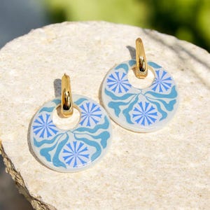 May include: A pair of gold-tone hoop earrings with circular, white pendants. Each pendant features a blue floral design. The earrings are displayed on a light-colored, textured surface. The earrings are a fashion accessory.