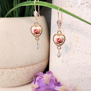 May include: Gold heart-shaped earrings with a floral design and clear crystal accents. The earrings are dangling and have a delicate look.