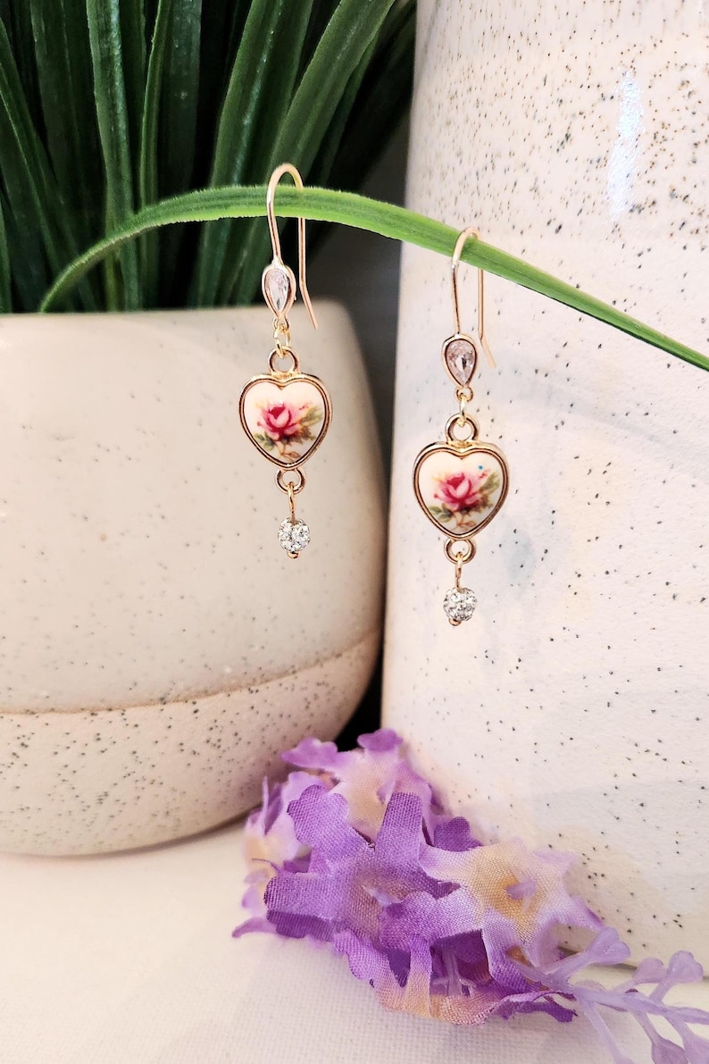 May include: Gold heart-shaped earrings with a floral design and clear crystal accents. The earrings are dangling and have a delicate look.