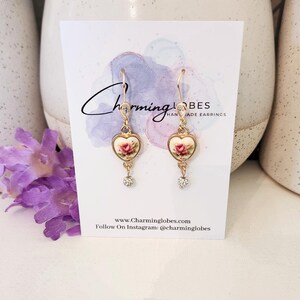 May include: A pair of gold heart-shaped earrings with a floral design and a clear crystal dangle. The earrings are on a white card with the text "Charming Lobes Handmade Earrings" and the website address "www.Charminglobes.com".