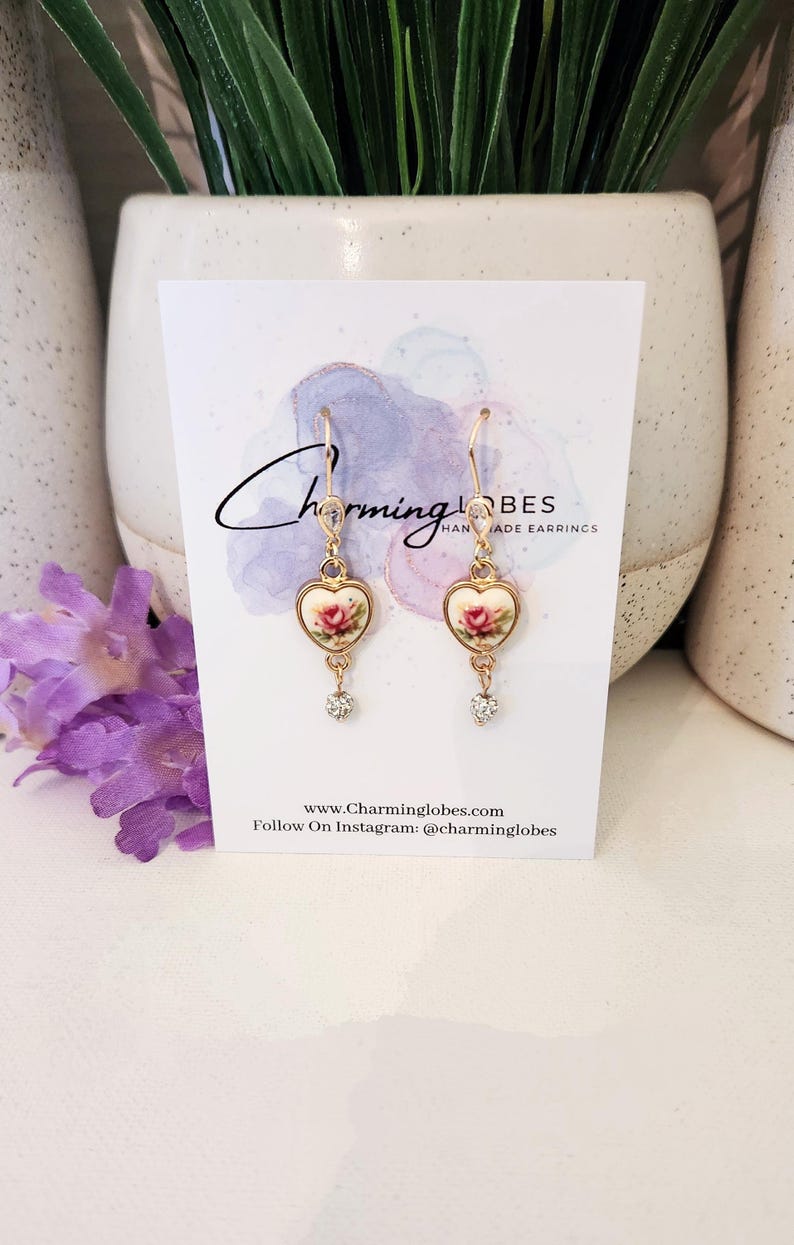 May include: A pair of gold heart-shaped earrings with a floral design and a clear crystal dangle. The earrings are on a white card with the text "Charming Lobes Handmade Earrings" and the website address "www.Charminglobes.com".