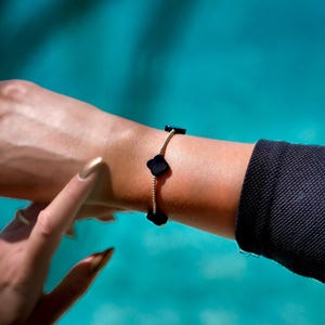 May include: A gold-toned bracelet with black clover-shaped charms. The bracelet is worn on a wrist with a gold-painted fingernail. The background is a blurred turquoise color.