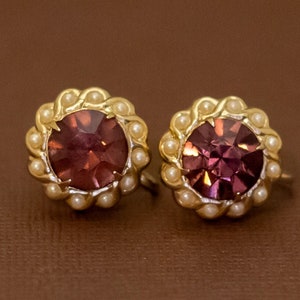 Könnte beinhalten: Ein Paar Vintage-Ohrstecker. Jeder Ohrring hat einen großen, runden, tiefvioletten Edelstein, der von kleinen, cremefarbenen Kunstperlen umgeben ist. Die Fassung ist goldfarben. Die Ohrringe werden auf einer braunen Oberfläche präsentiert.