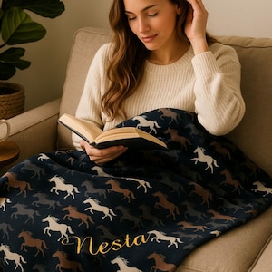 Könnte beinhalten: Eine marineblaue Decke mit einem sich wiederholenden Muster aus weißen, braunen und grauen Pferden. Der Name "Nesta" ist in goldener Schrift gestickt. Die Person sitzt in einem beigen Sessel und liest ein Buch.