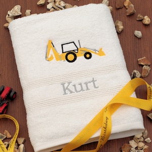 May include: White bath towel with a yellow excavator embroidered on it. The name "Kurt" is embroidered below the excavator. A yellow ribbon with the text "Happy Birthday" is wrapped around the towel.