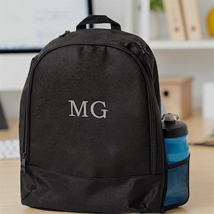 Può includere: Zaino nero con le iniziali "MG" in lettere argentate. Lo zaino ha uno scomparto principale con cerniera e una tasca laterale che contiene una bottiglia d'acqua blu. La borsa ha una maniglia superiore e una cerniera frontale.