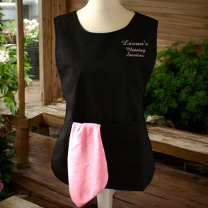 Puede incluir: Delantal negro sin mangas con un pa&ntilde;o de limpieza rosa en el bolsillo. El delantal tiene el texto "Lauren's Cleaning Services" bordado en rosa en la parte delantera.