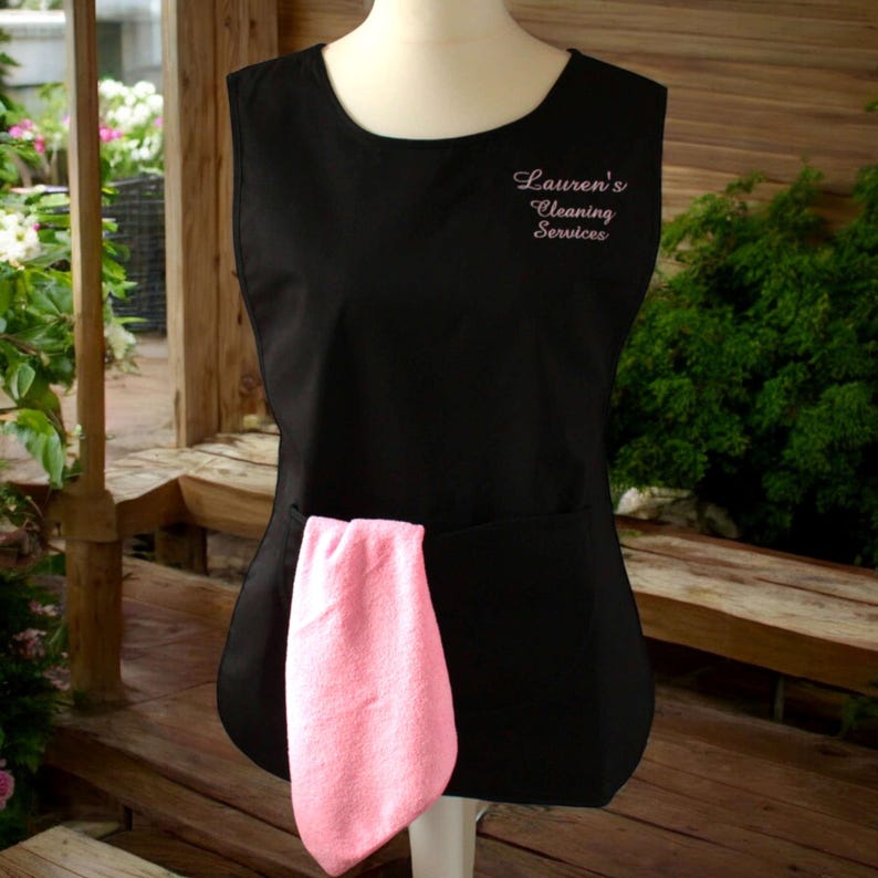 Puede incluir: Delantal negro sin mangas con un pa&ntilde;o de limpieza rosa en el bolsillo. El delantal tiene el texto "Lauren's Cleaning Services" bordado en rosa en la parte delantera.