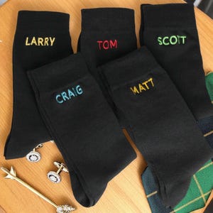 May include: Five pairs of black socks with different names embroidered on them in various colors. The names are Larry, Tom, Scott, Craig, and Matt.