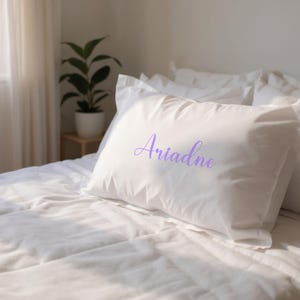 May include: White pillowcase with the name "Ariadne" in a lavender script. The pillow is on a bed with white bedding. A potted plant is visible in the background.