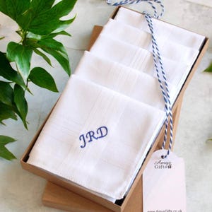May include: A set of white cotton handkerchiefs folded in a brown cardboard box. The top handkerchief features the blue embroidered initials JRD. A blue and white striped string is tied around the box with a white tag labelled Amys Gifts.