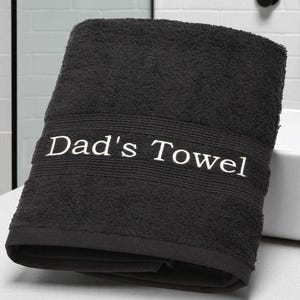May include: A dark grey bath towel with the embroidered text "Dad's Towel" in white. The towel is folded and has a textured surface, with a ribbed band where the text is displayed. The towel is likely made of absorbent material.